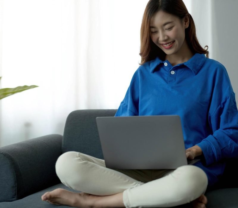 Young business freelance Asian woman working on laptop checking social media while lying on the sofa when relax in living room at home.